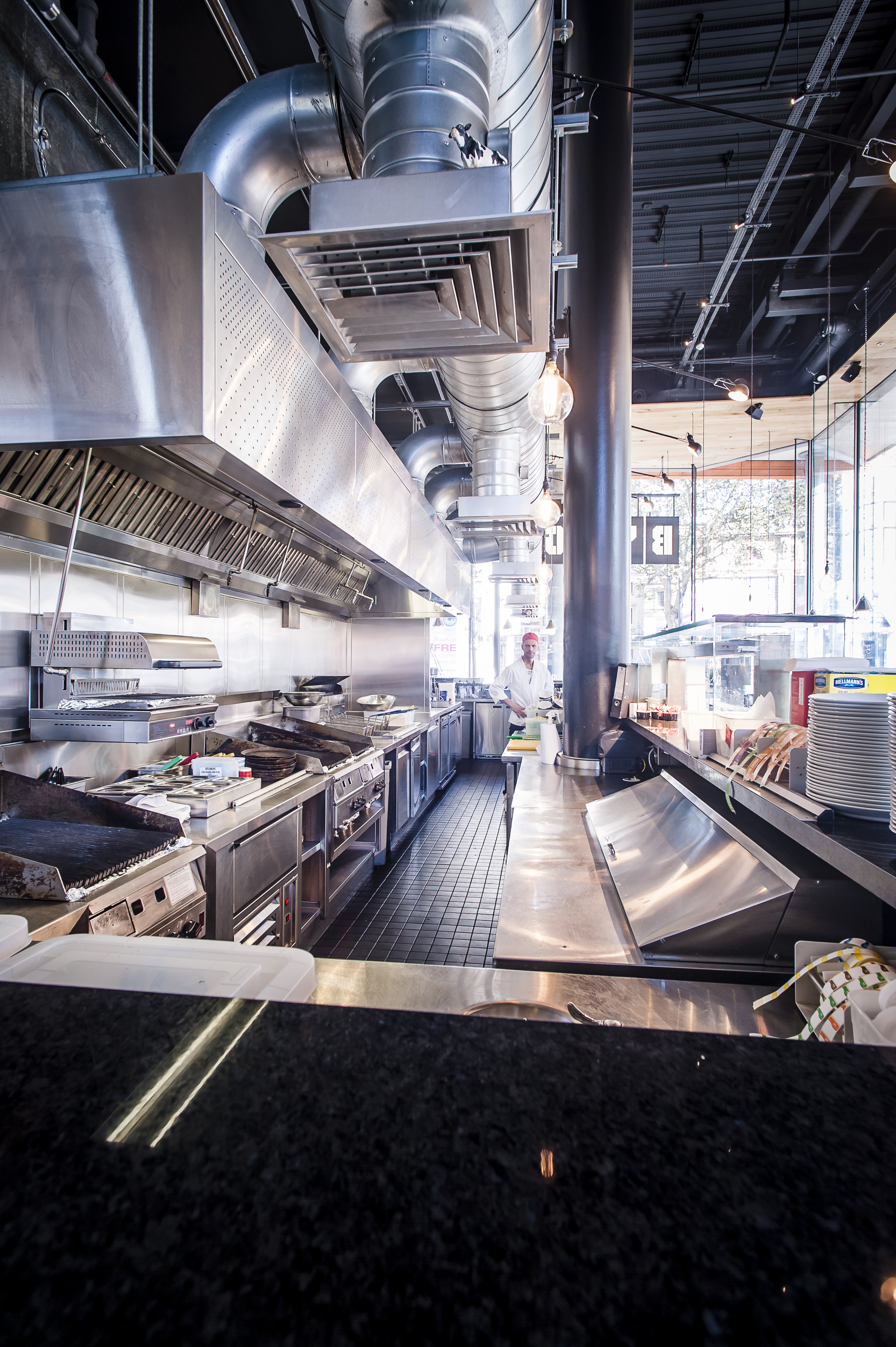 Photo shows the space behind the counter in a commercial kitchen with a big extractor fan and ducting above the grill and fryers